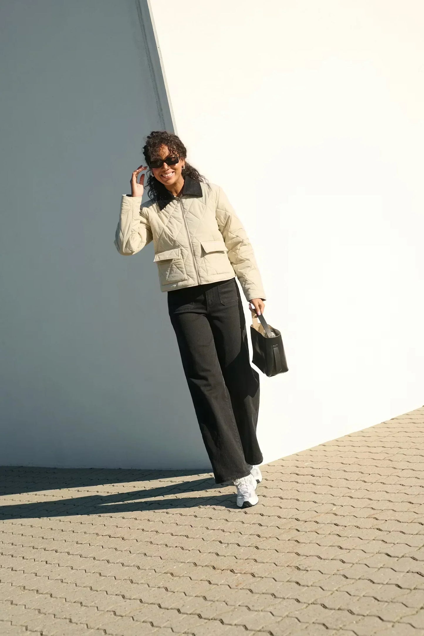 Woman in quilted beige jacket, black pants, and white sneakers posing outdoors with handbag
