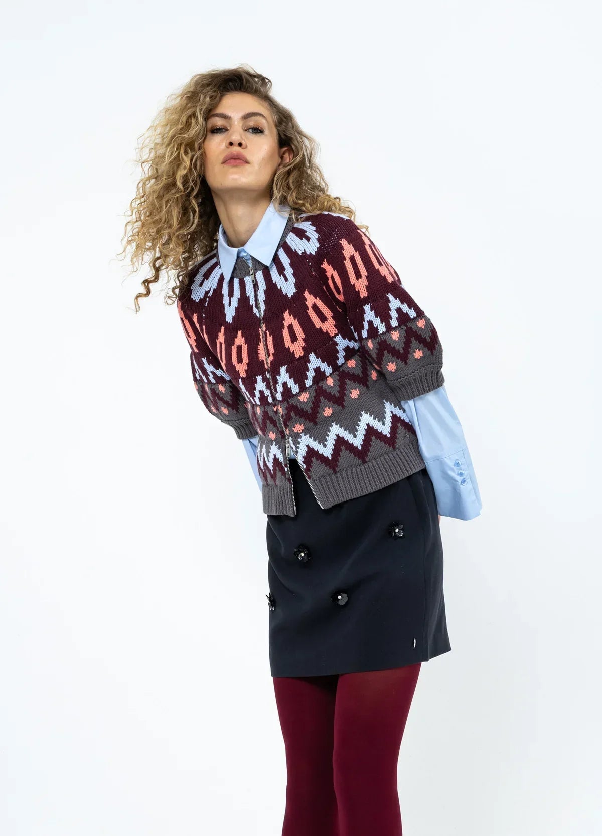 Woman in patterned knit sweater, collared shirt, black skirt with flower accents, and burgundy tights