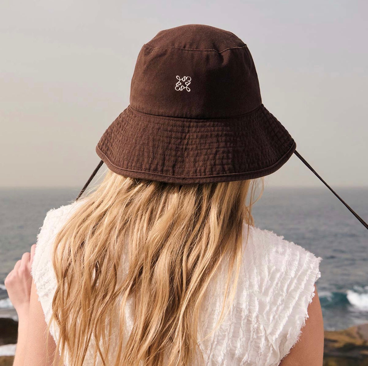 Brown bucket hat with white logo, worn by woman with blonde hair overlooking ocean, coastal fashion