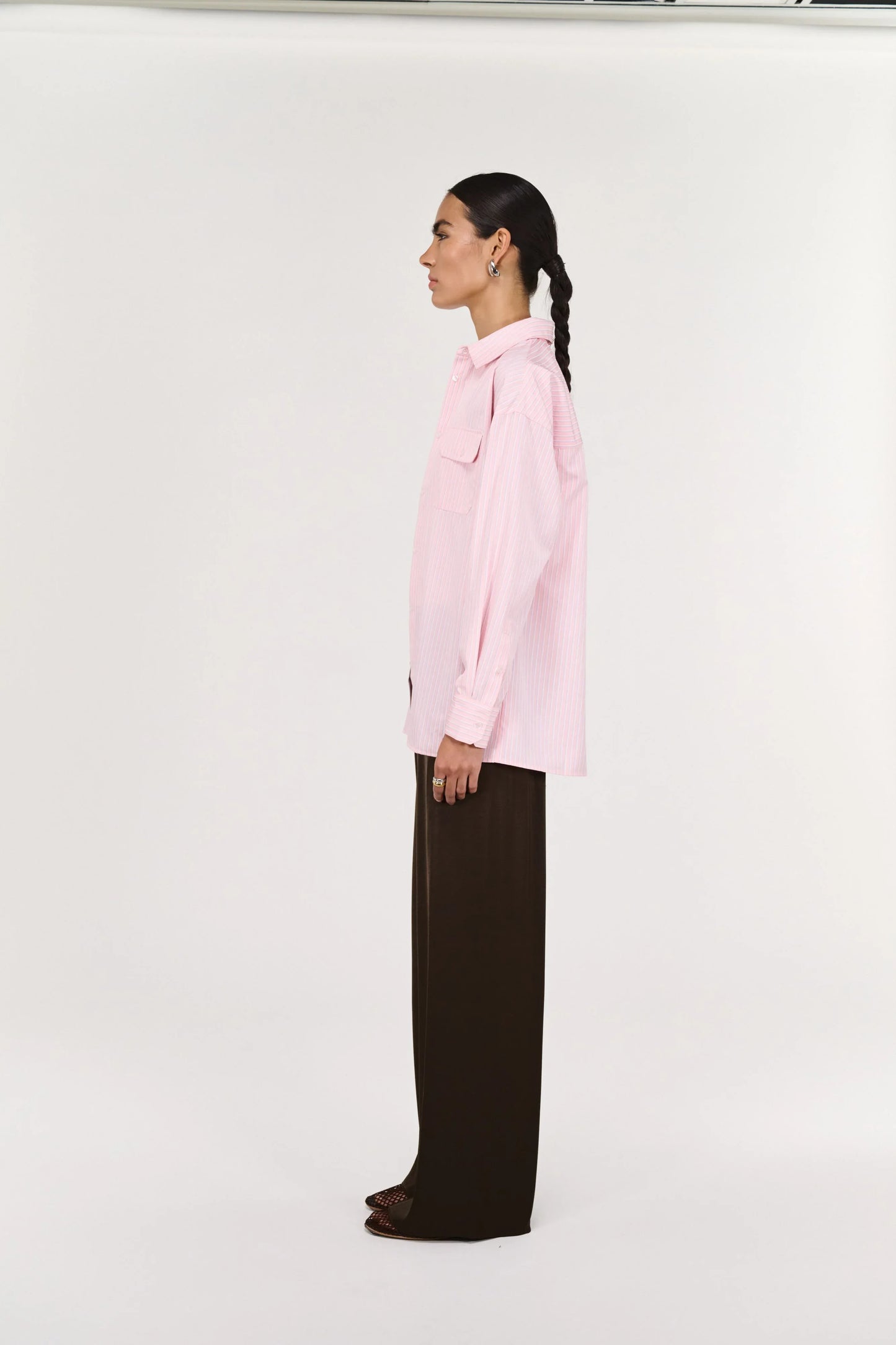 Woman in a pink striped oversized shirt and brown wide-leg pants, side profile, studio background
