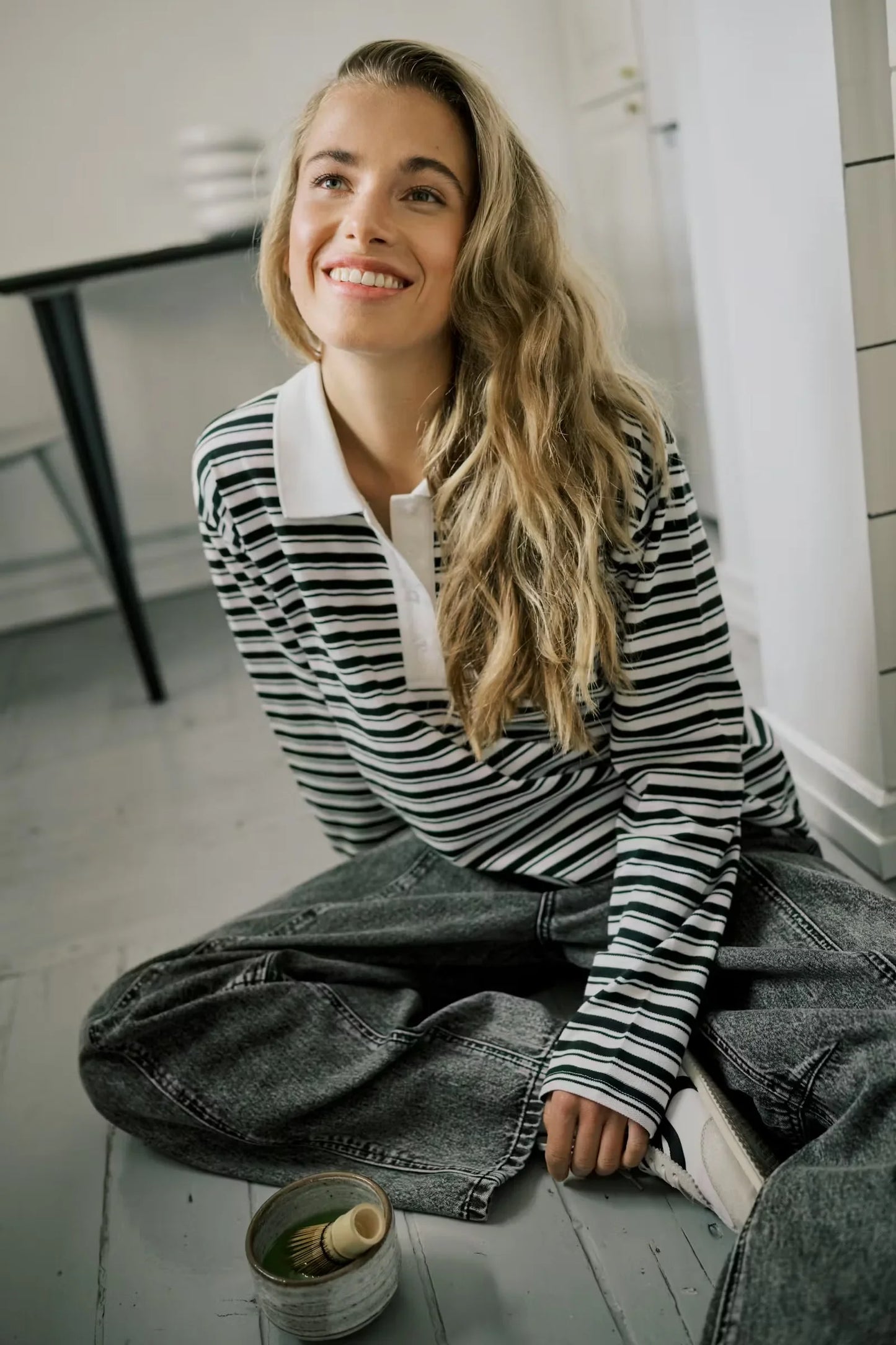 Woman in striped long sleeve polo shirt and grey jeans, sitting on floor with matcha bowl
