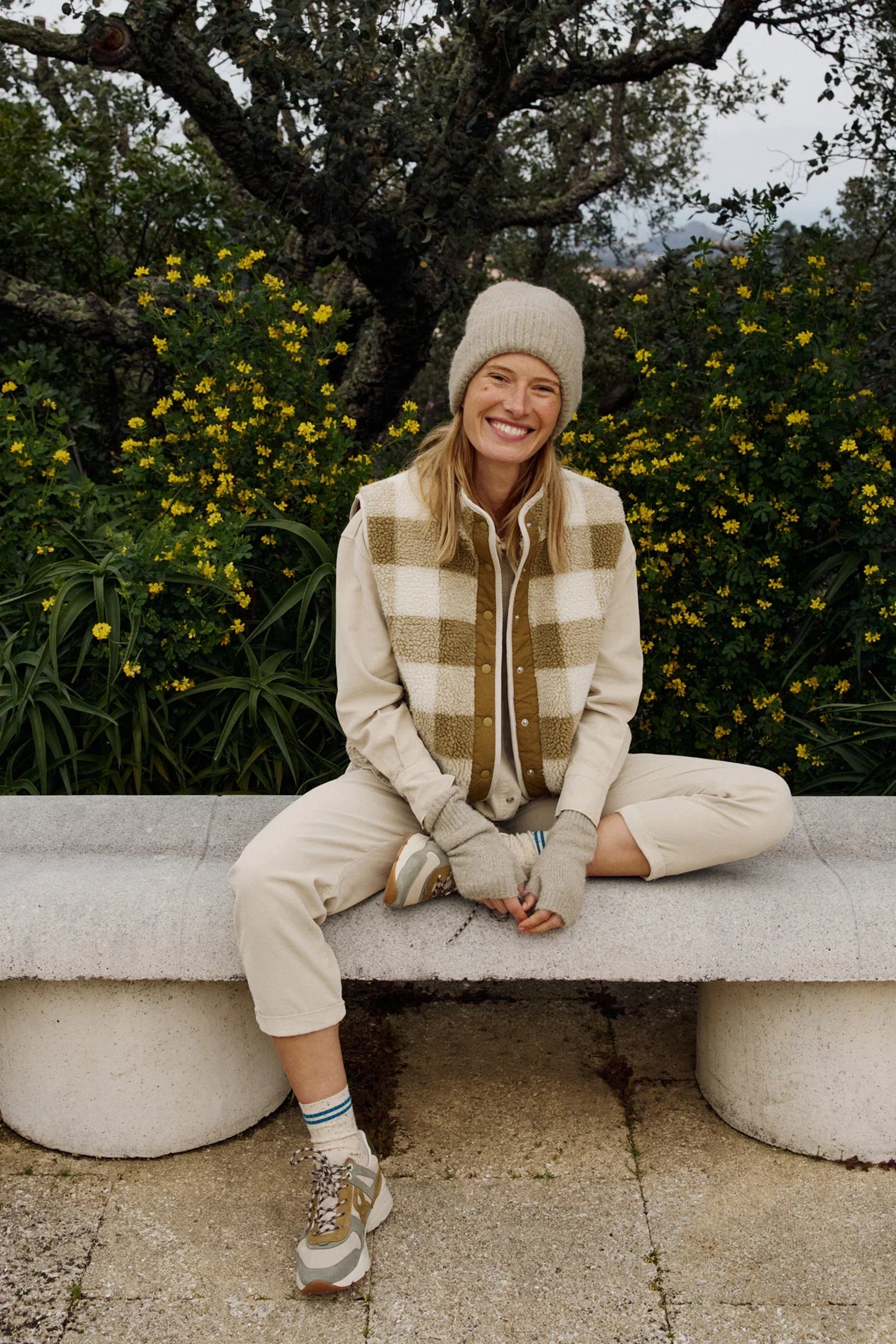 Woman in cozy plaid vest, beanie, and sneakers sitting on bench outdoors with yellow flowers