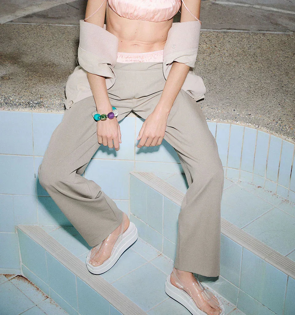 Woman in beige wide-leg pants and clear platform sandals sitting on tiled steps, wearing colorful beaded bracelet