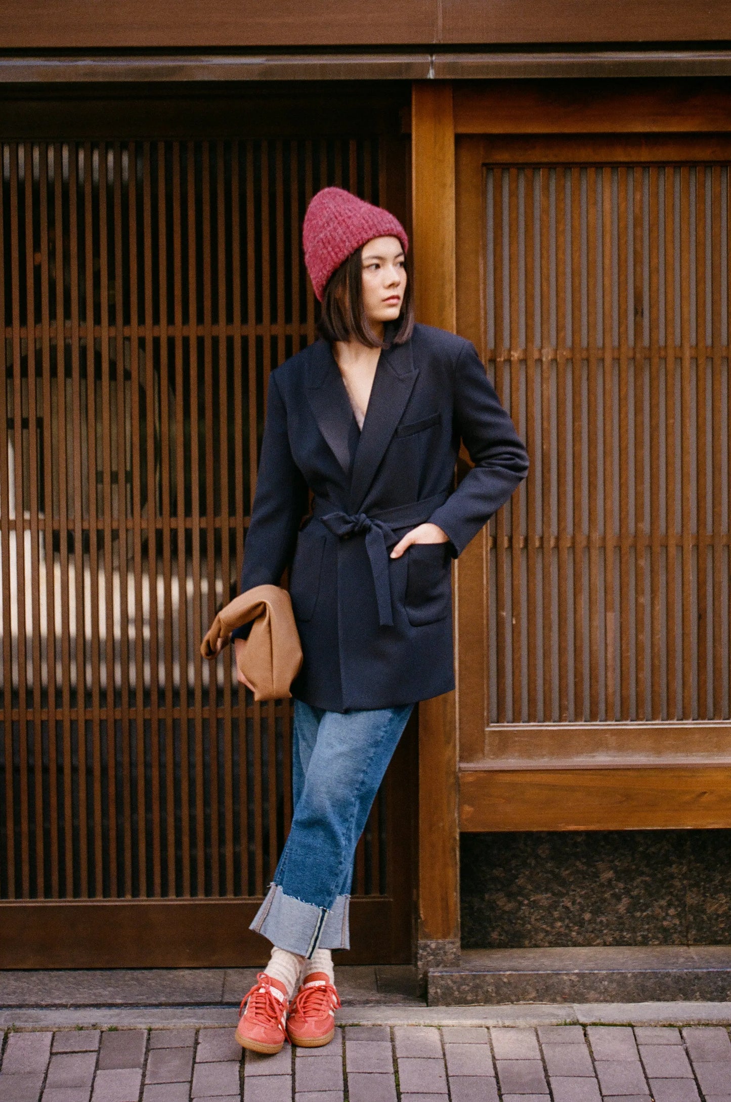 Woman in navy belted blazer, red beanie, and cuffed jeans with orange sneakers, urban street style