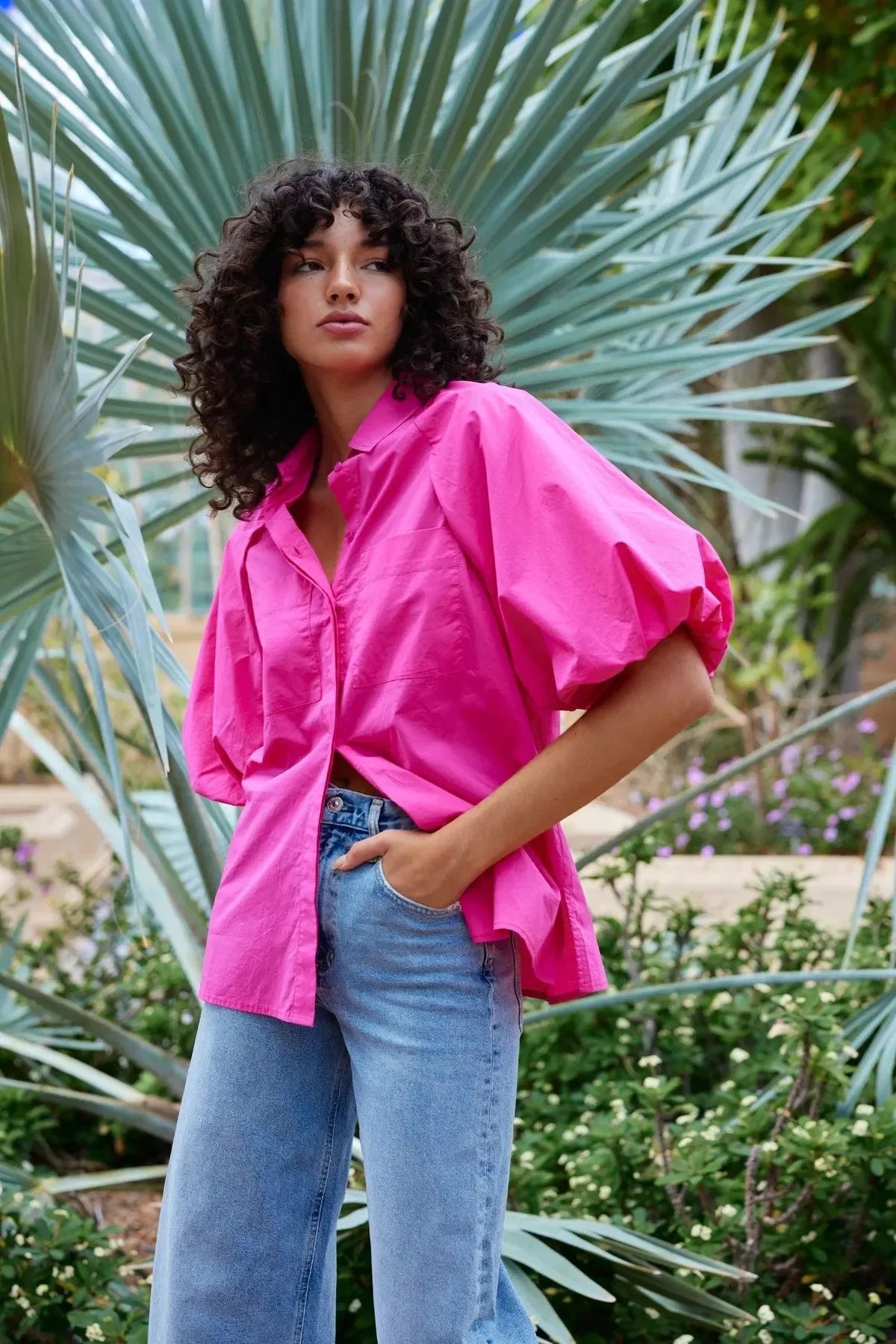 Woman in vibrant pink oversized shirt with puff sleeves, paired with blue wide-leg jeans, styled for bold everyday fashion.