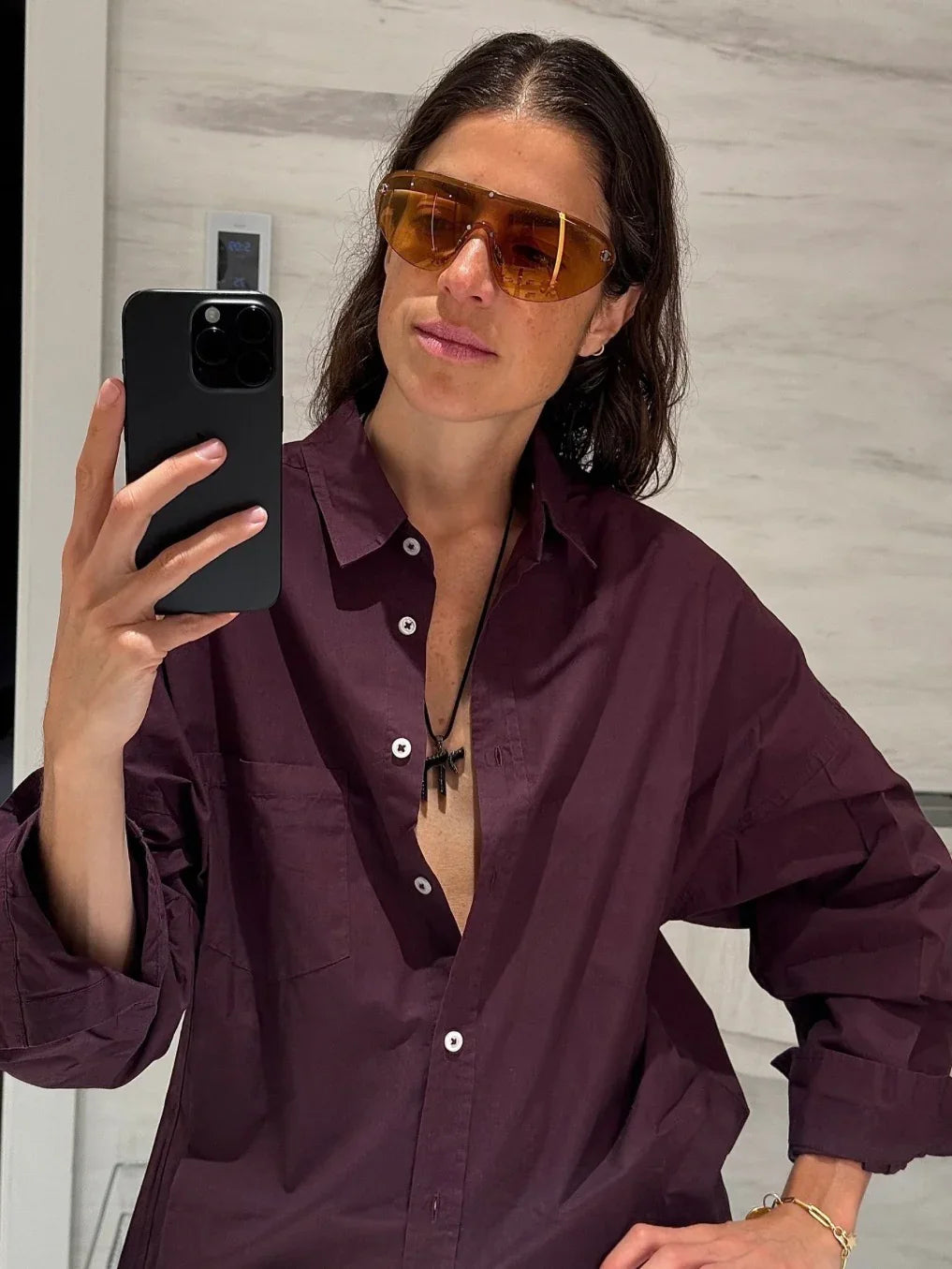 Woman in maroon shirt and amber sunglasses taking mirror selfie with black phone