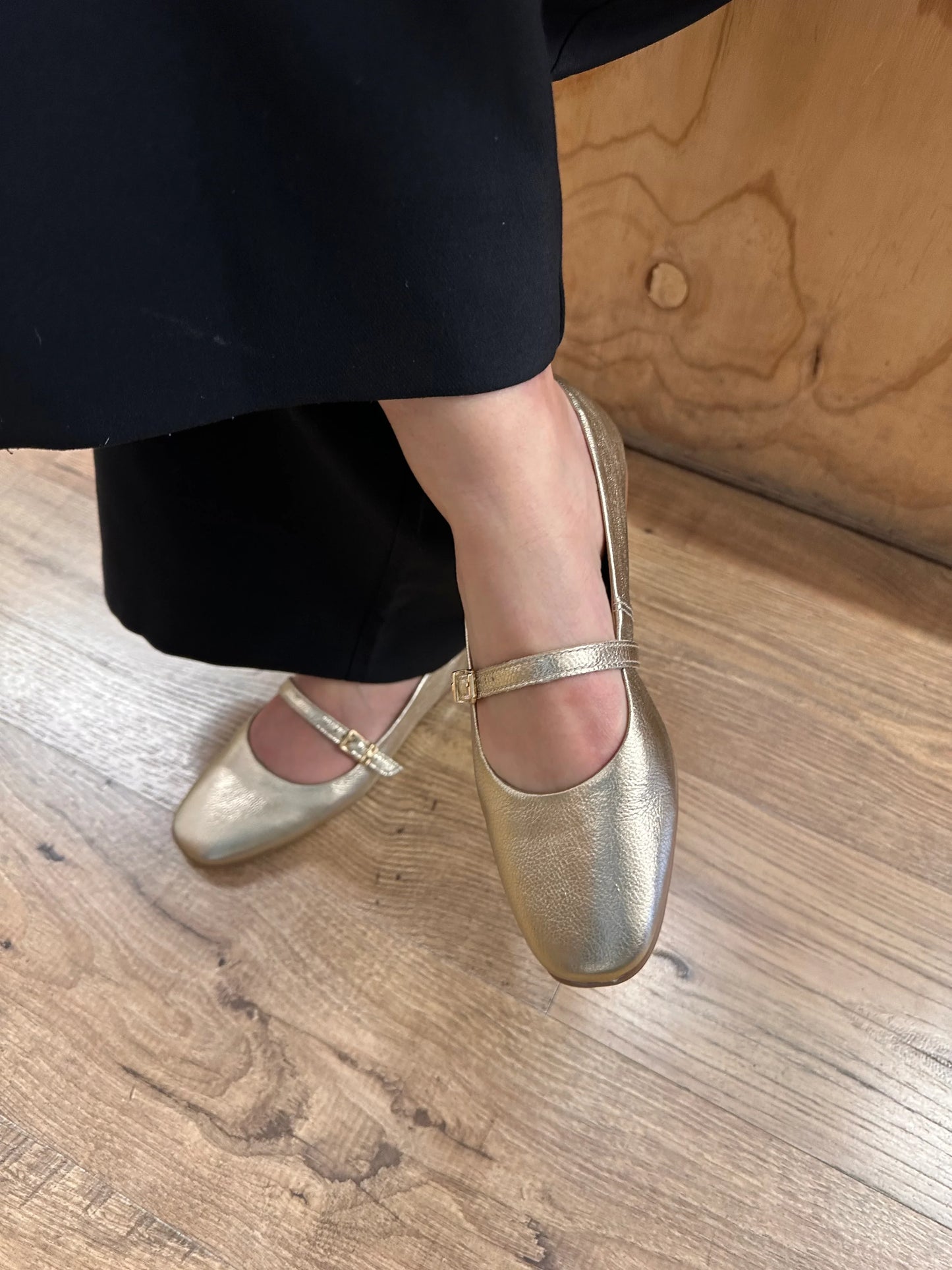 Gold Mary Jane flats with buckle strap, paired with black trousers on wooden floor