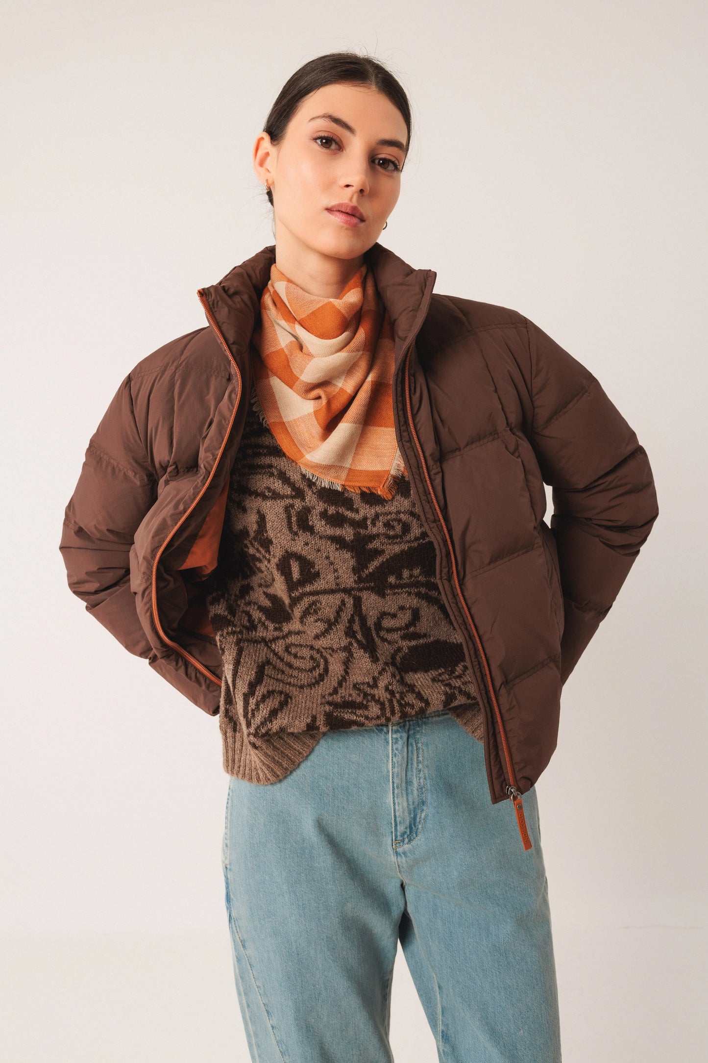 Woman in brown puffer jacket, patterned sweater, orange scarf, and blue jeans