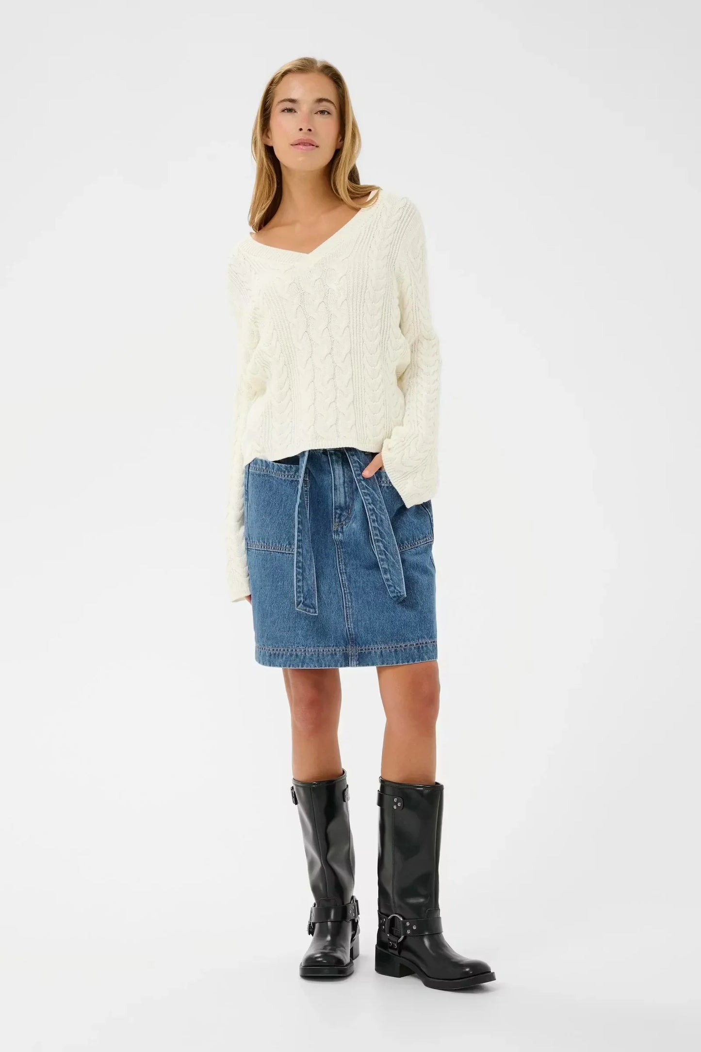 Woman in cream cable knit sweater, denim skirt with tie waist, and black buckle boots