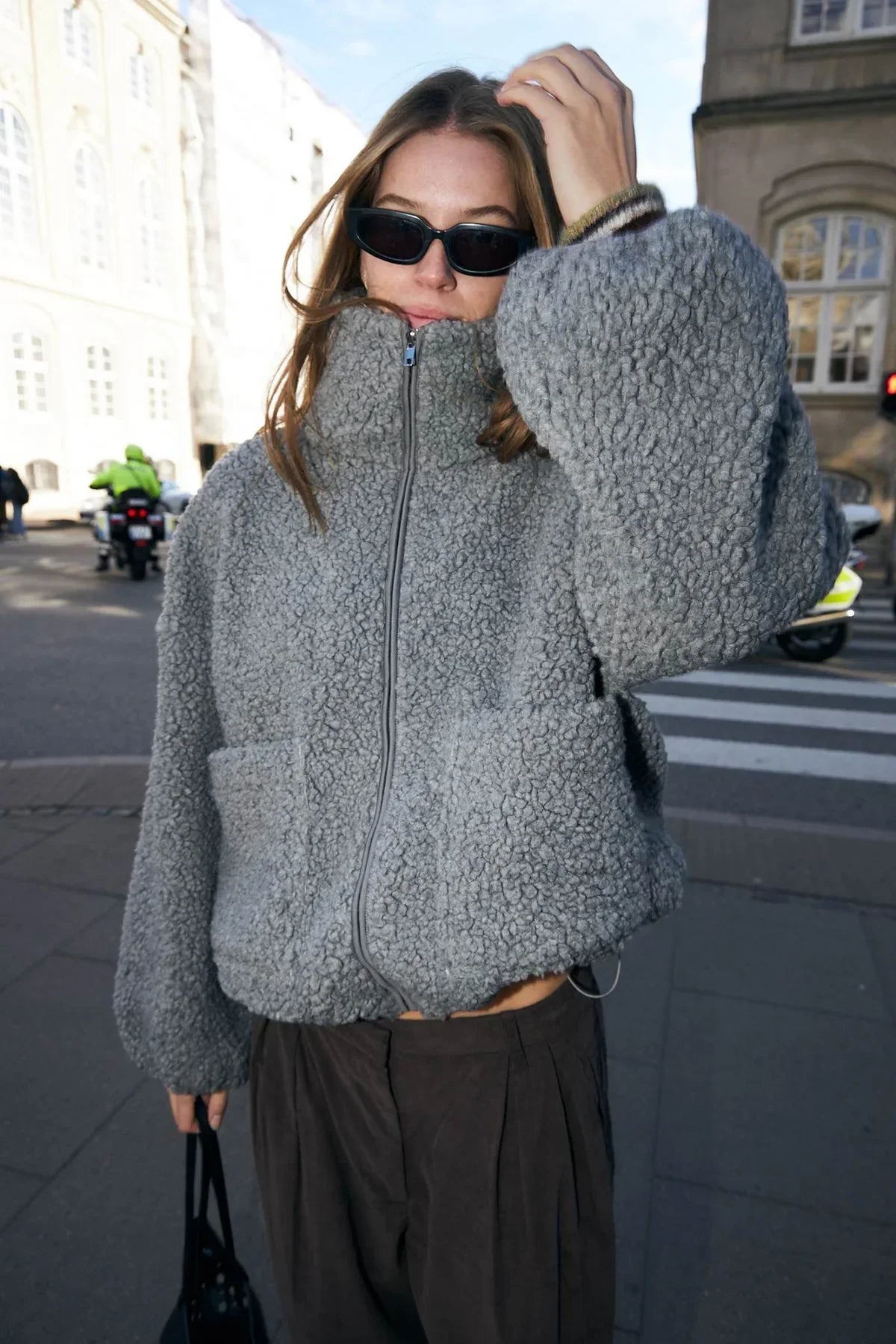 Woman wearing oversized grey sherpa jacket, sunglasses, and pleated brown pants on a city street