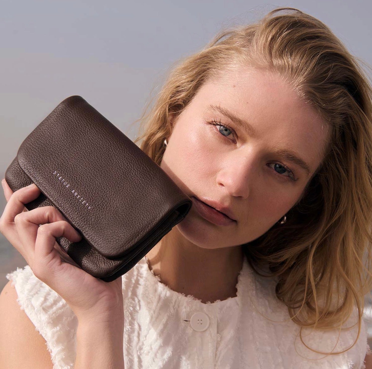 Woman holding brown leather wallet with 'STATUS ANXIETY' embossed, styled with white textured top