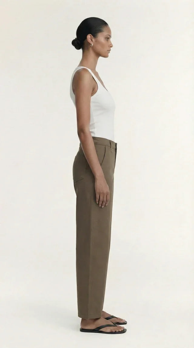 Woman in a white ribbed tank top and olive green tailored trousers standing in profile.