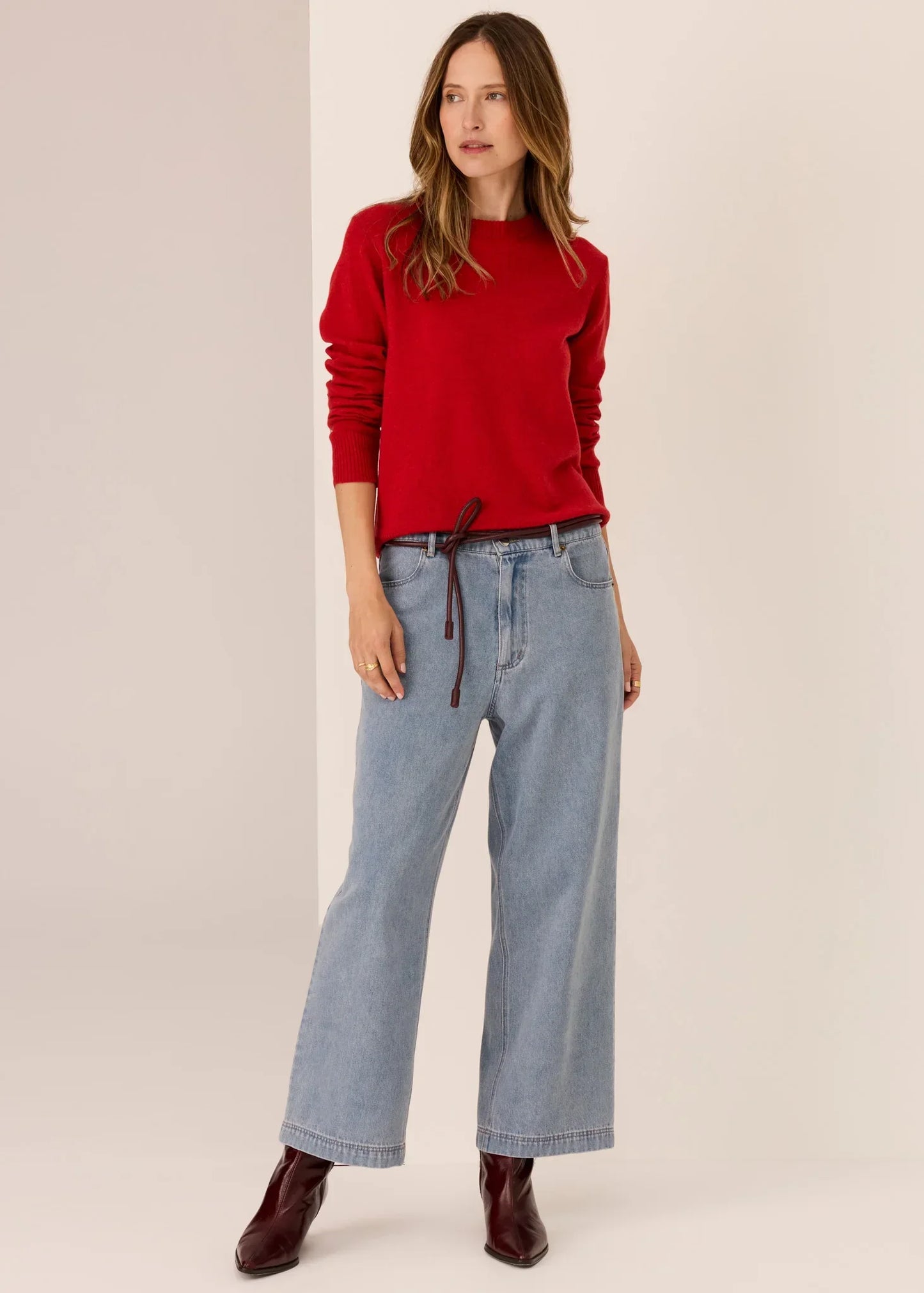 Woman in red sweater, wide-leg light blue jeans, and burgundy boots on neutral background
