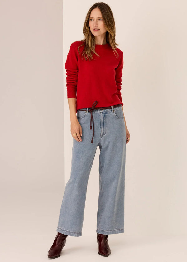 Woman in red sweater, wide-leg light blue jeans, and burgundy boots on neutral background
