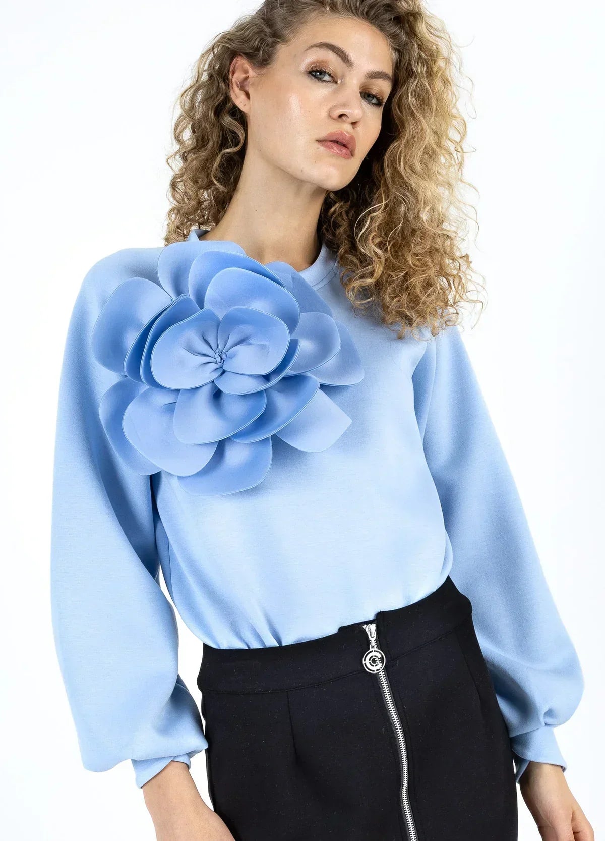 Woman wearing light blue blouse with large 3D floral applique and black zip skirt