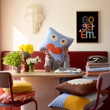 Person holding a blue character face decorative pillow in a bright living room setting.