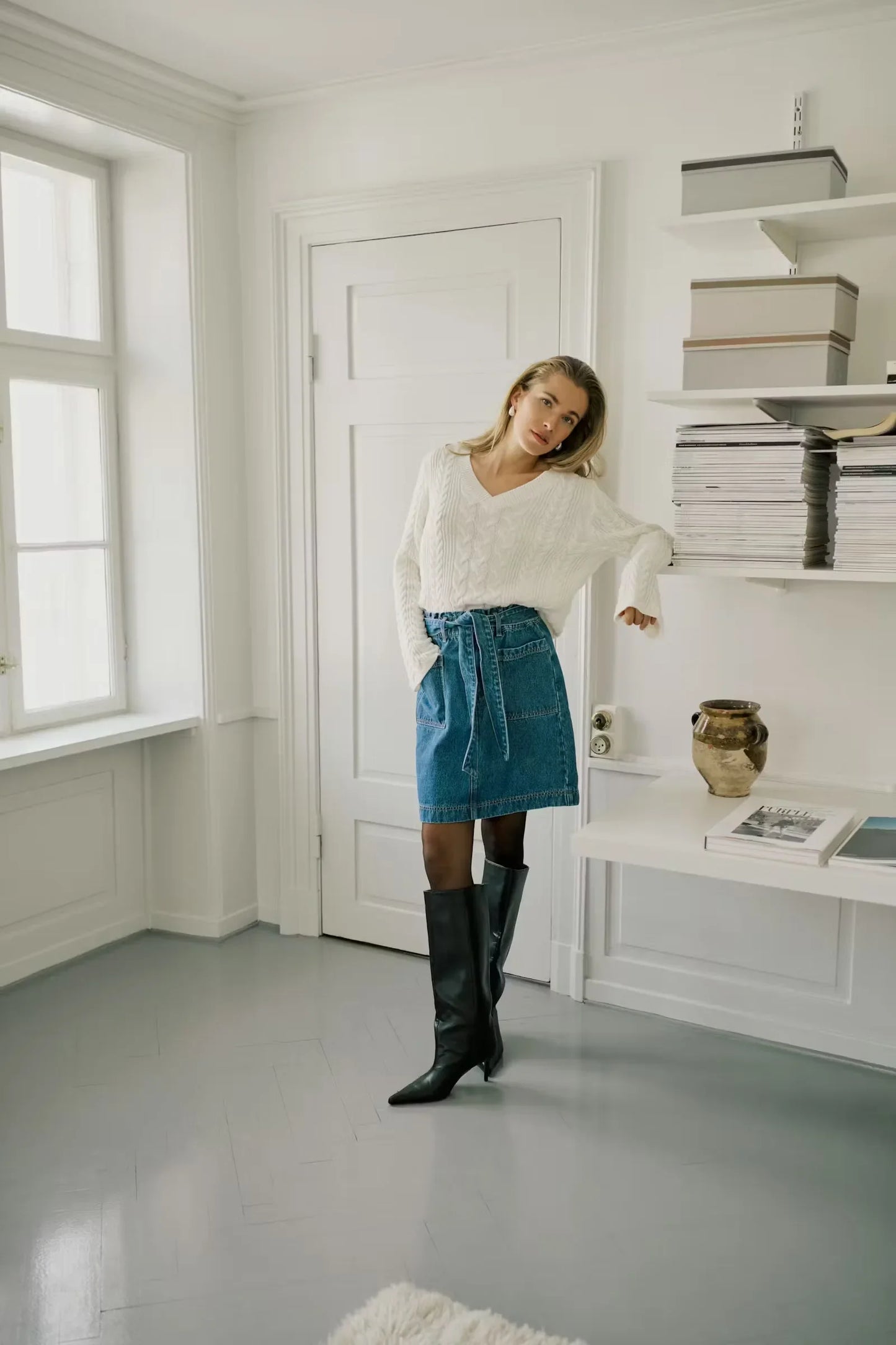 Woman in white cable knit sweater, blue denim wrap skirt, and black knee-high boots in bright room