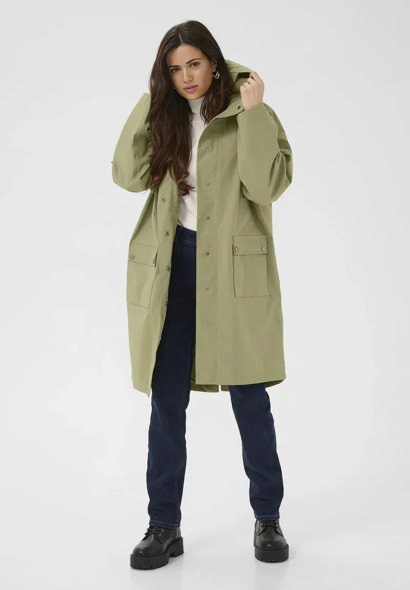 Olive green women's raincoat with hood, front pockets, and button closure, styled with jeans and boots