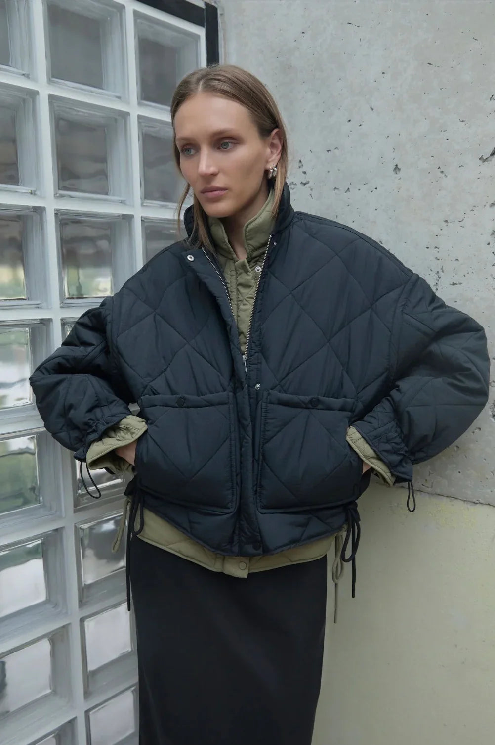 Woman wearing black quilted puffer jacket with olive green lining, hands in pockets, standing by glass block wall