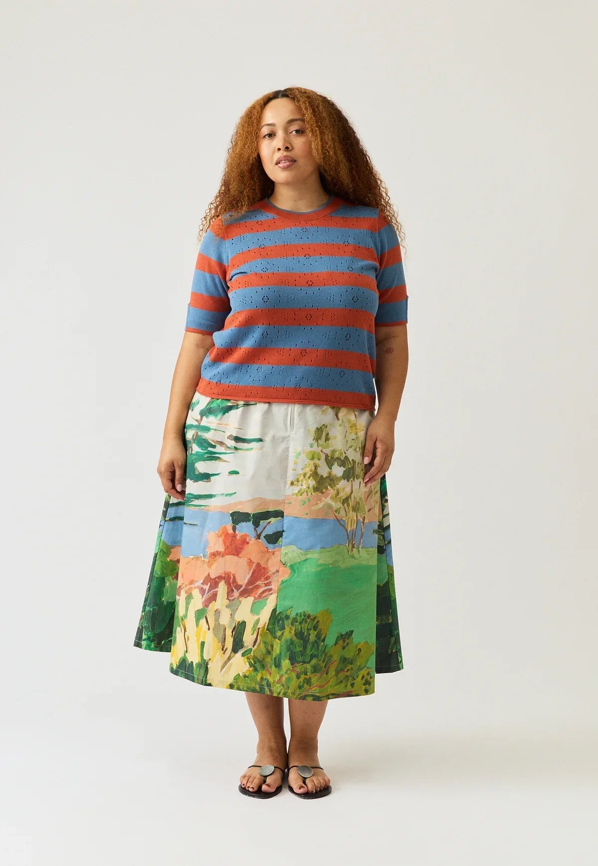 Woman wearing NANCYBIRD Rana Knit Tee in Terracotta Stripe and a landscape print skirt.