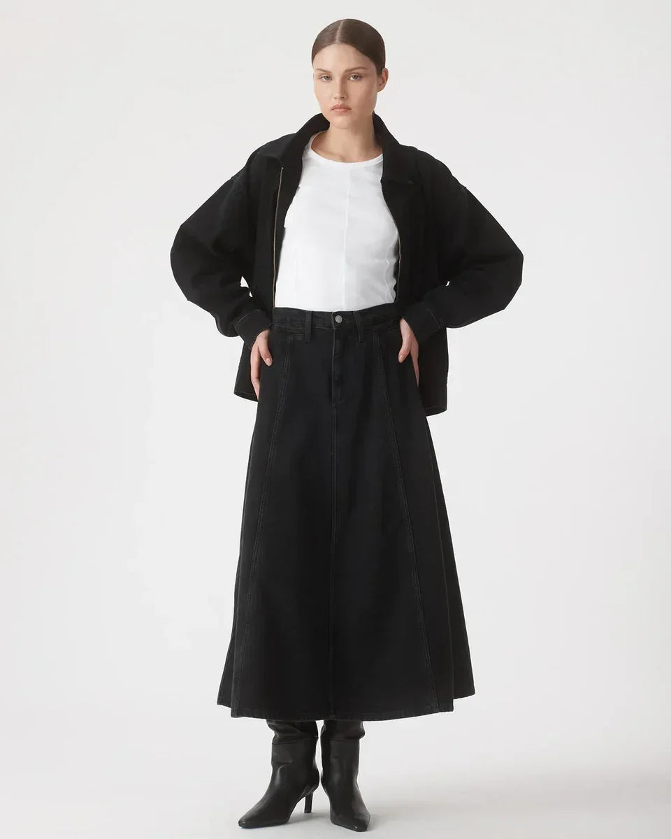 Woman in black denim midi skirt, white top, black jacket, and heeled boots, styled for modern women's fashion.