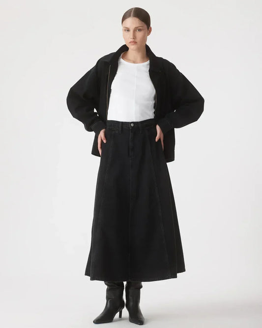 Woman in black denim midi skirt, white top, black jacket, and heeled boots, styled for modern women's fashion.