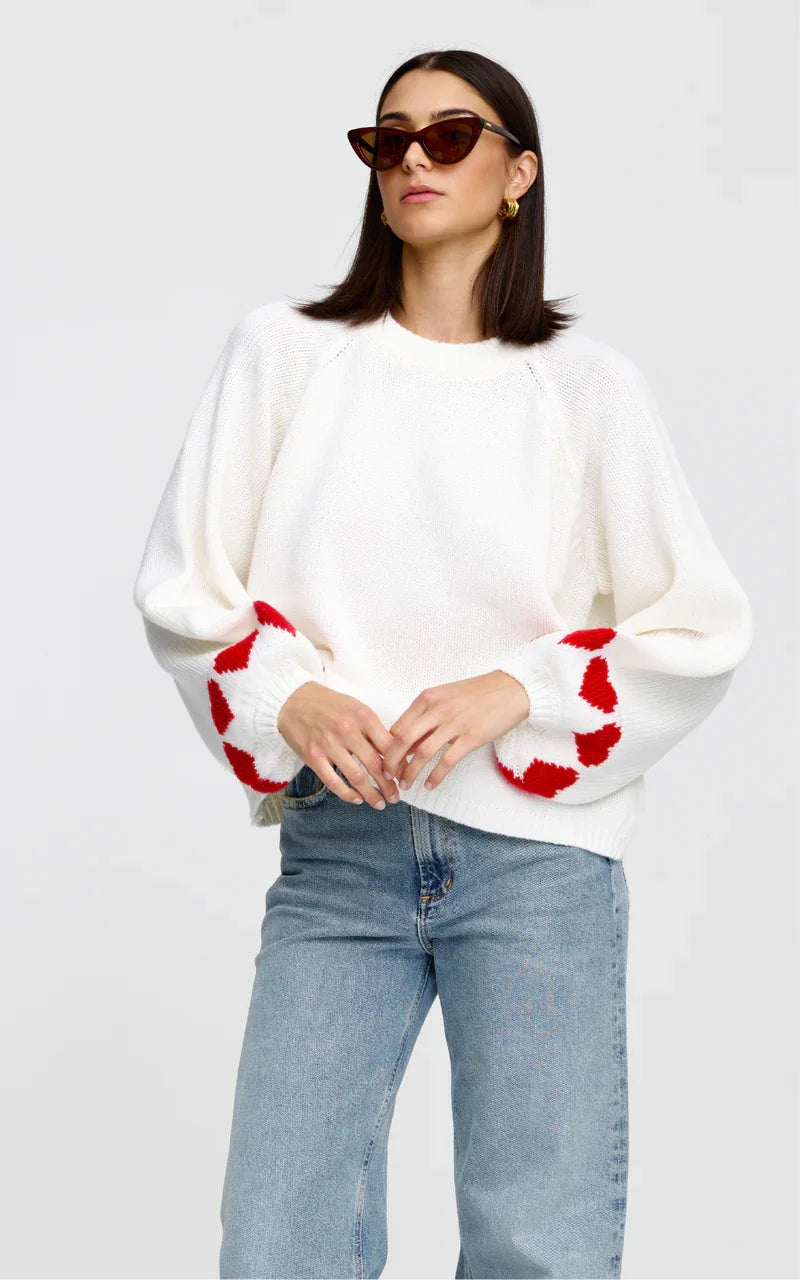 White knit sweater with red heart pattern on sleeves, paired with blue jeans and sunglasses