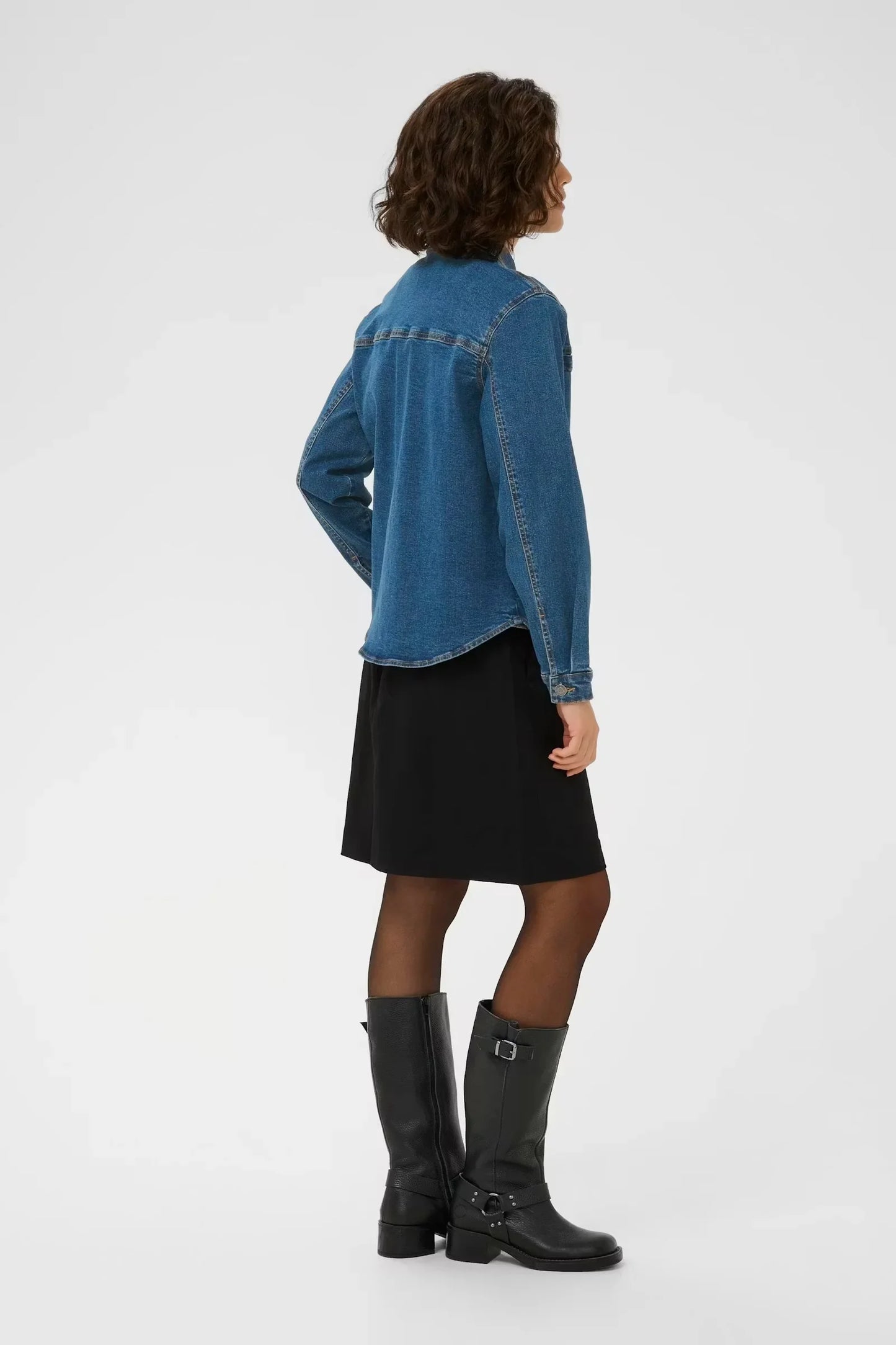 Woman in blue denim shirt, black skirt, and knee-high boots, side view