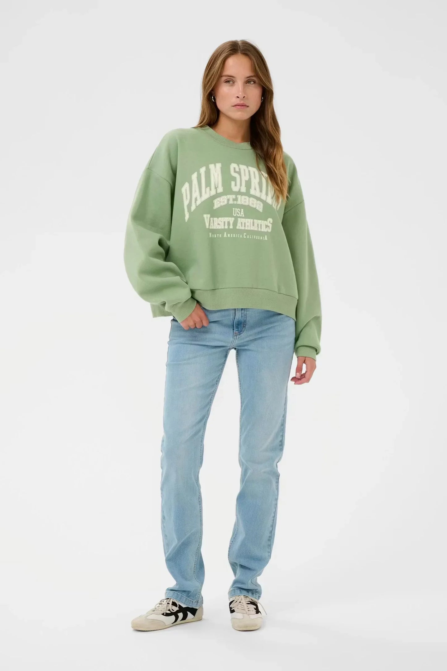 Woman in green Palm Springs varsity sweatshirt with light wash jeans and sneakers