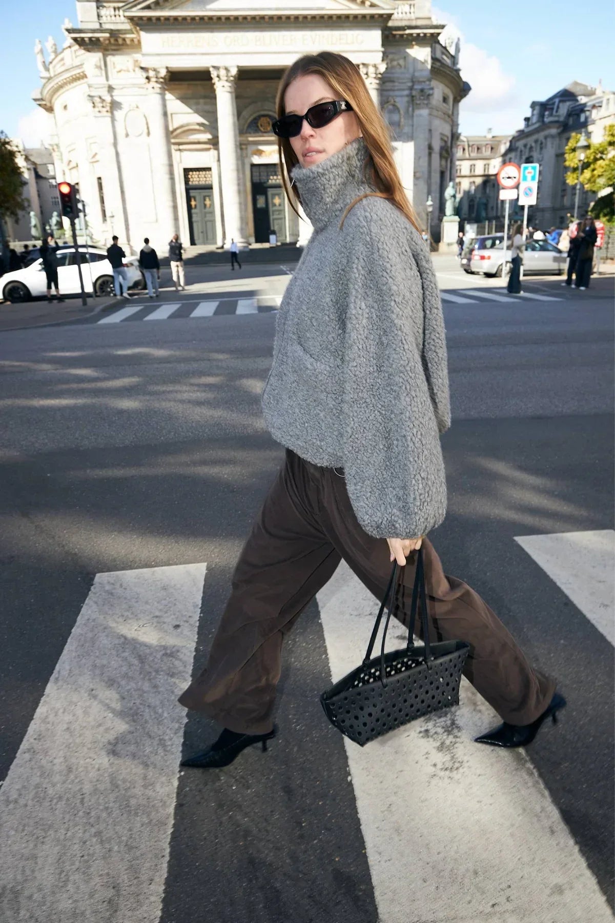 Woman in grey shearling jacket, brown wide-leg pants, and black heeled shoes crossing street with a black tote bag