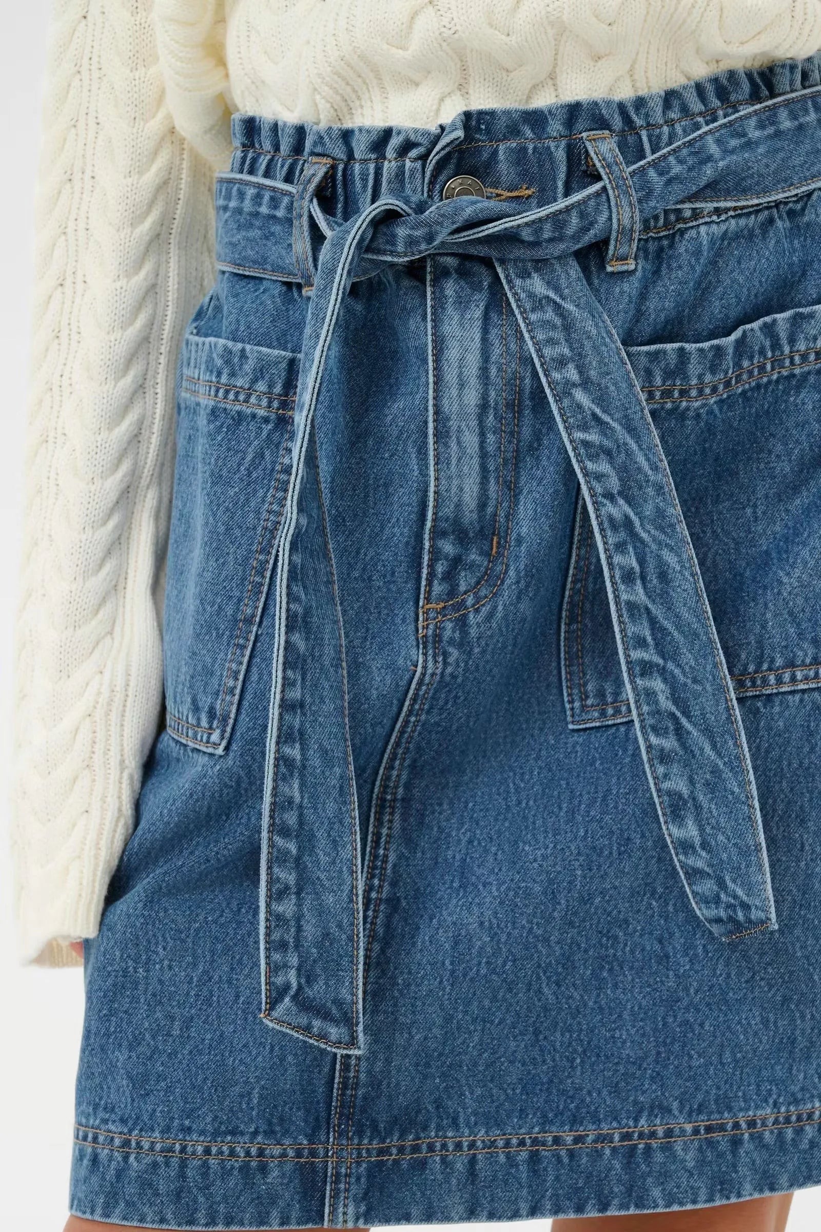 Blue denim mini skirt with tie waist and front pockets, paired with cream cable knit sweater