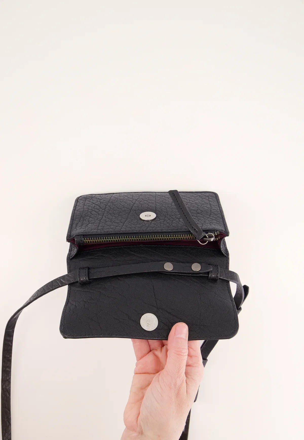 Hand holding open black leather crossbody bag with snap closure and zipper compartment