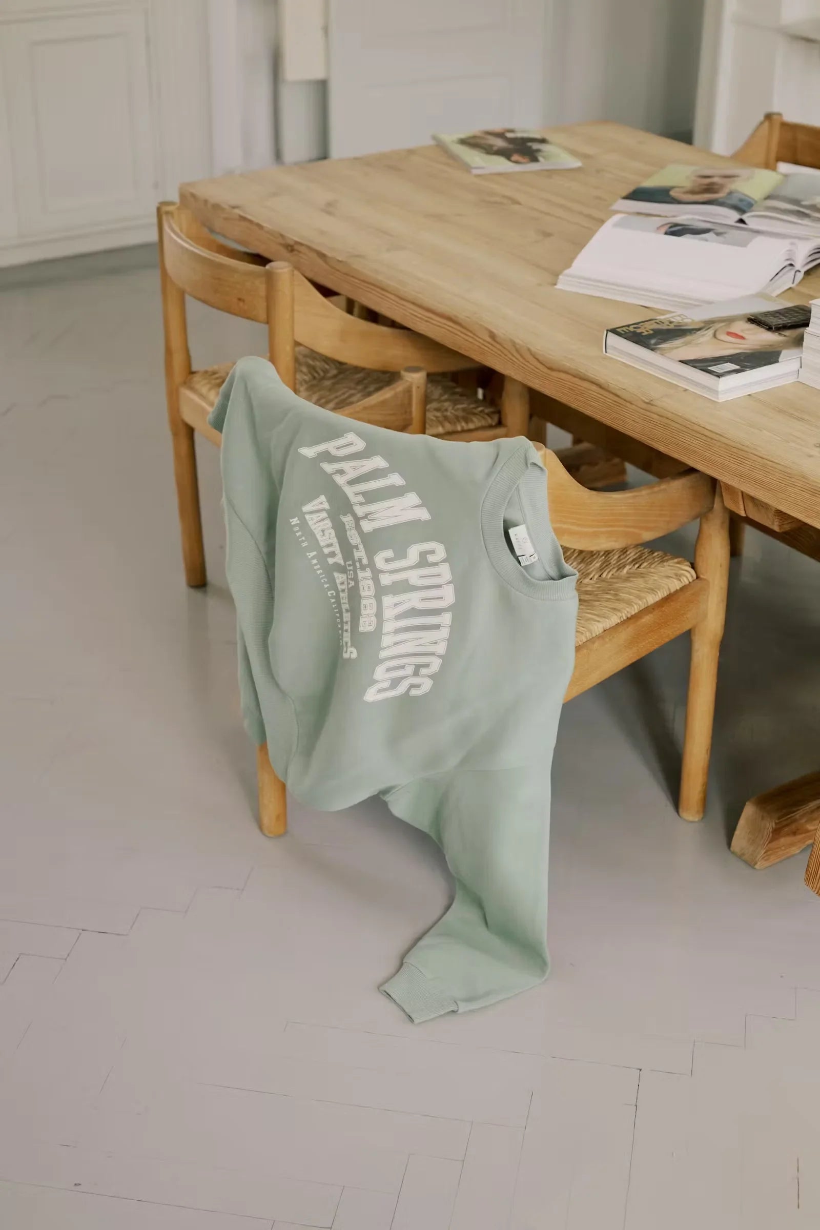 Mint green Palm Springs varsity sweatshirt draped on wooden chair near rustic table with magazines
