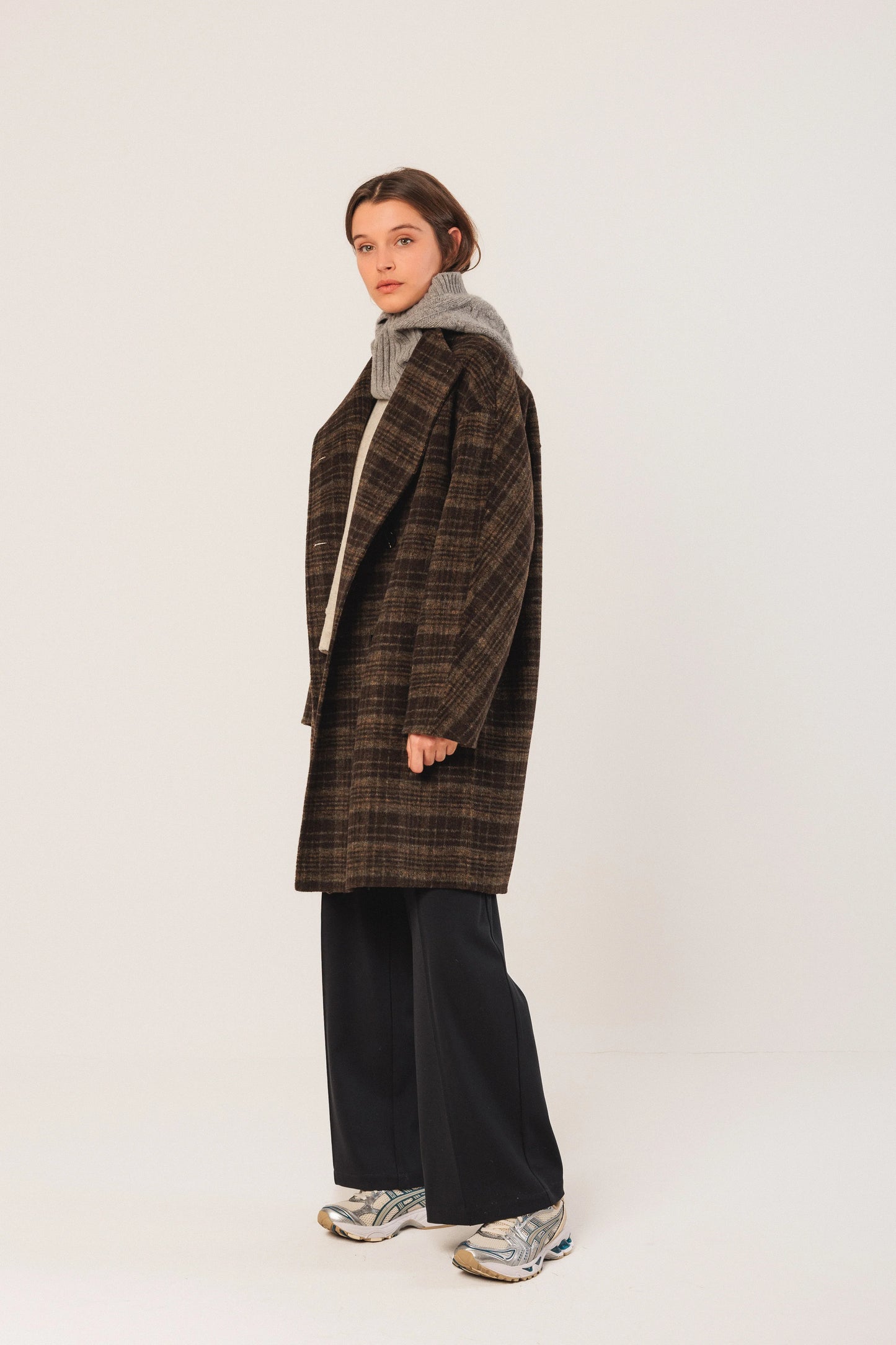 Woman in brown plaid coat, grey turtleneck, black wide-leg pants, and silver sneakers, studio look