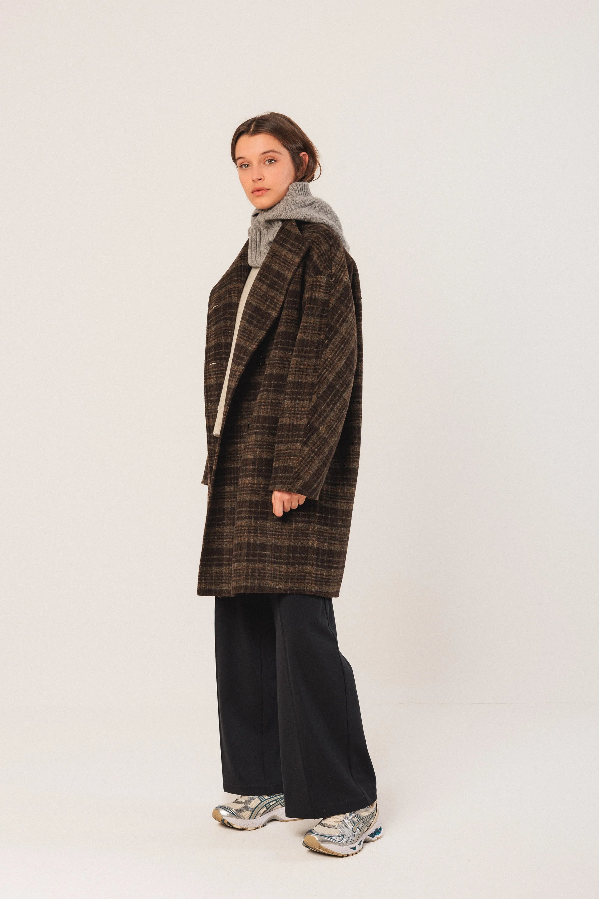 Woman in brown plaid coat, grey turtleneck, black wide-leg pants, and silver sneakers, studio look