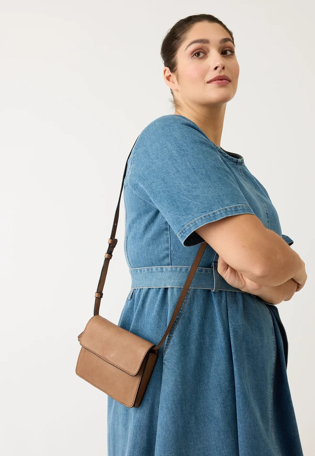 Woman wearing blue denim dress with tan crossbody bag, posing against white background