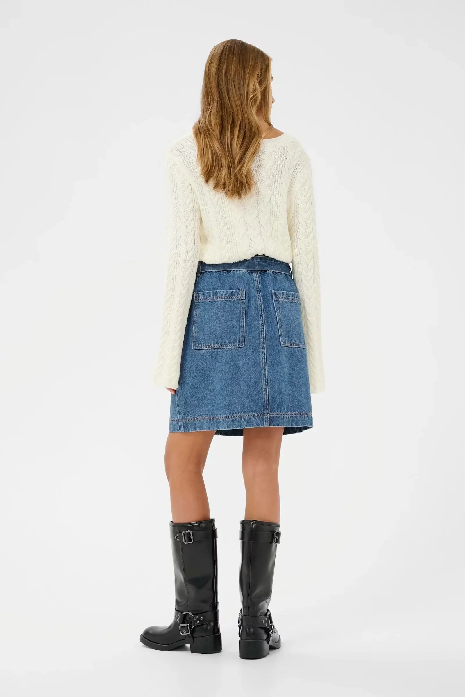 Woman in cream cable knit sweater, blue denim skirt with back pockets, and black buckle boots