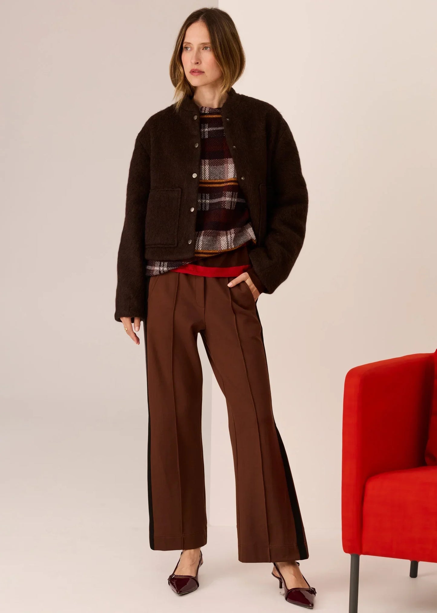 Woman in brown fuzzy jacket, plaid top, and wide-leg trousers with black side stripes, styled with burgundy heels.