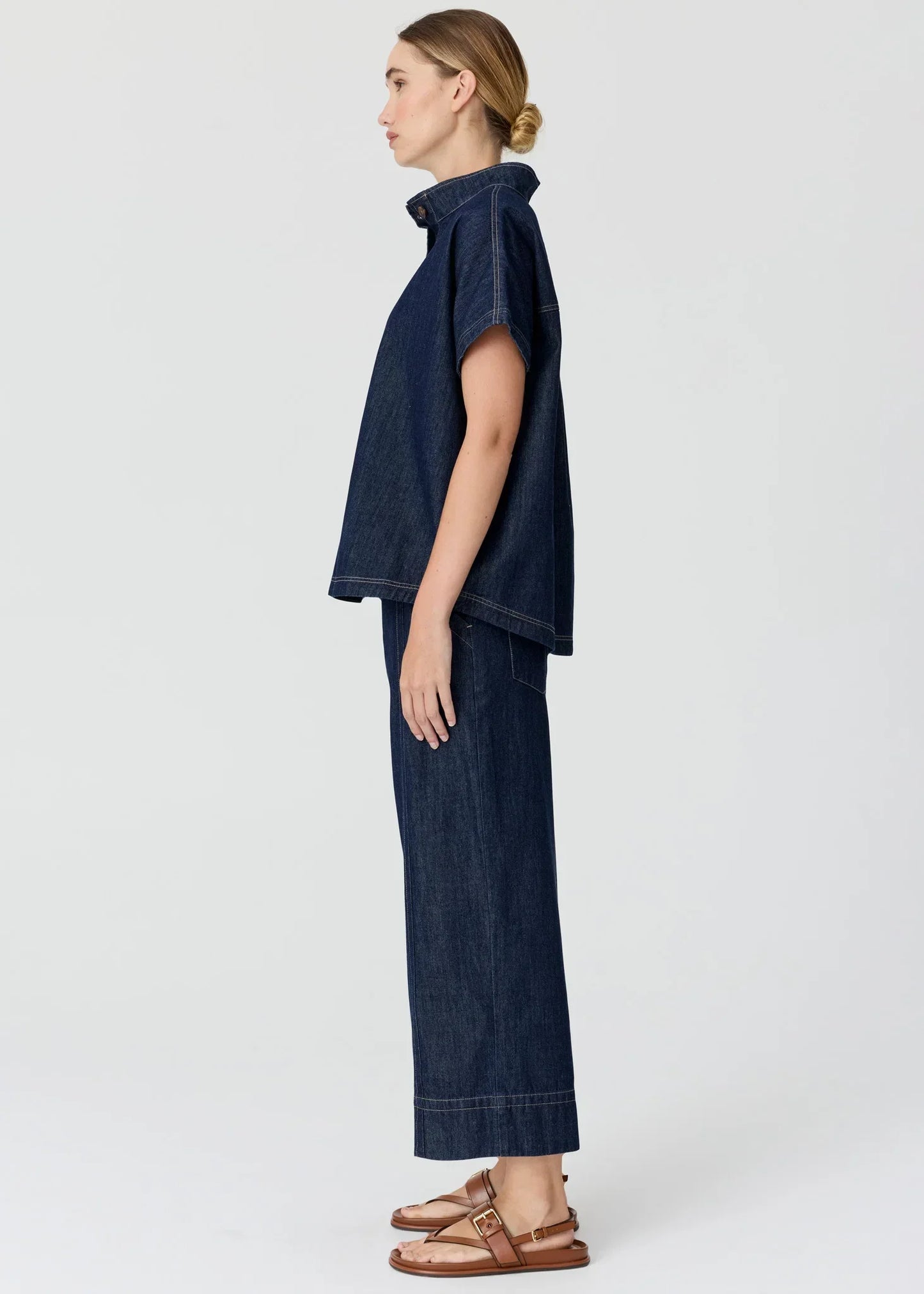 Woman in a dark blue denim short-sleeved shirt and wide-leg trousers with brown sandals.