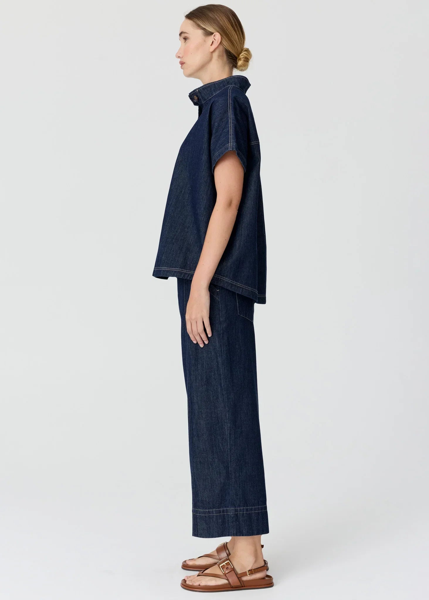 Woman in a dark blue denim short-sleeved shirt and wide-leg trousers with brown sandals.