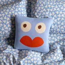 Blue whimsical face decorative pillow with large eyes and red lips on patterned bedding.