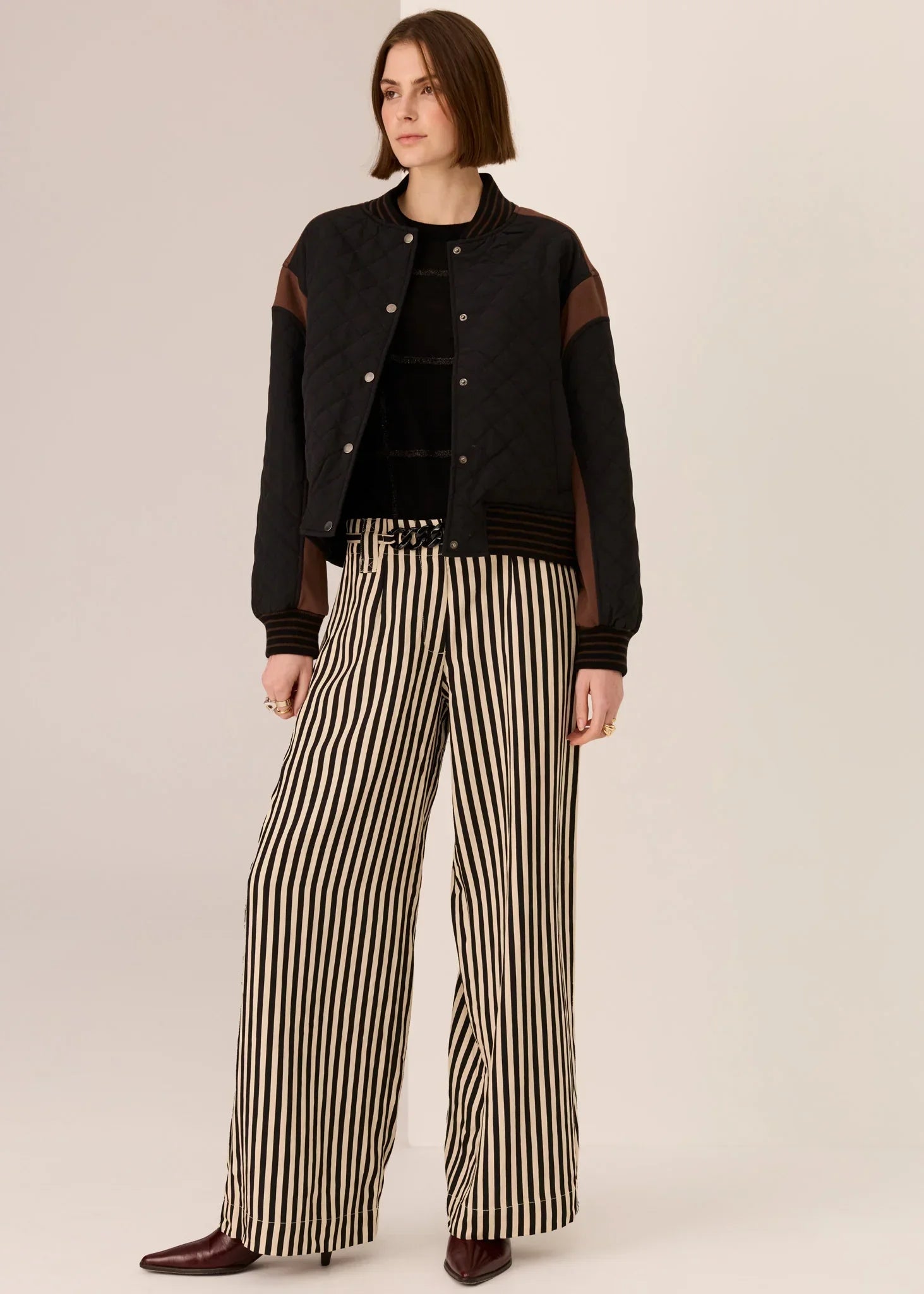 Woman wearing black quilted bomber jacket, striped wide-leg pants, and burgundy heels