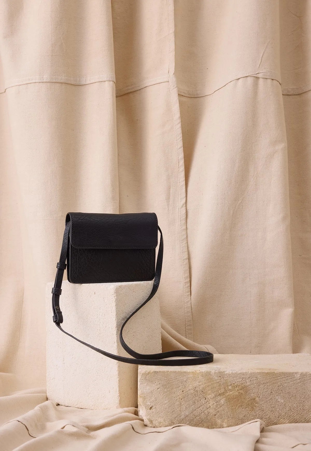 Black leather crossbody bag with adjustable strap on beige fabric backdrop