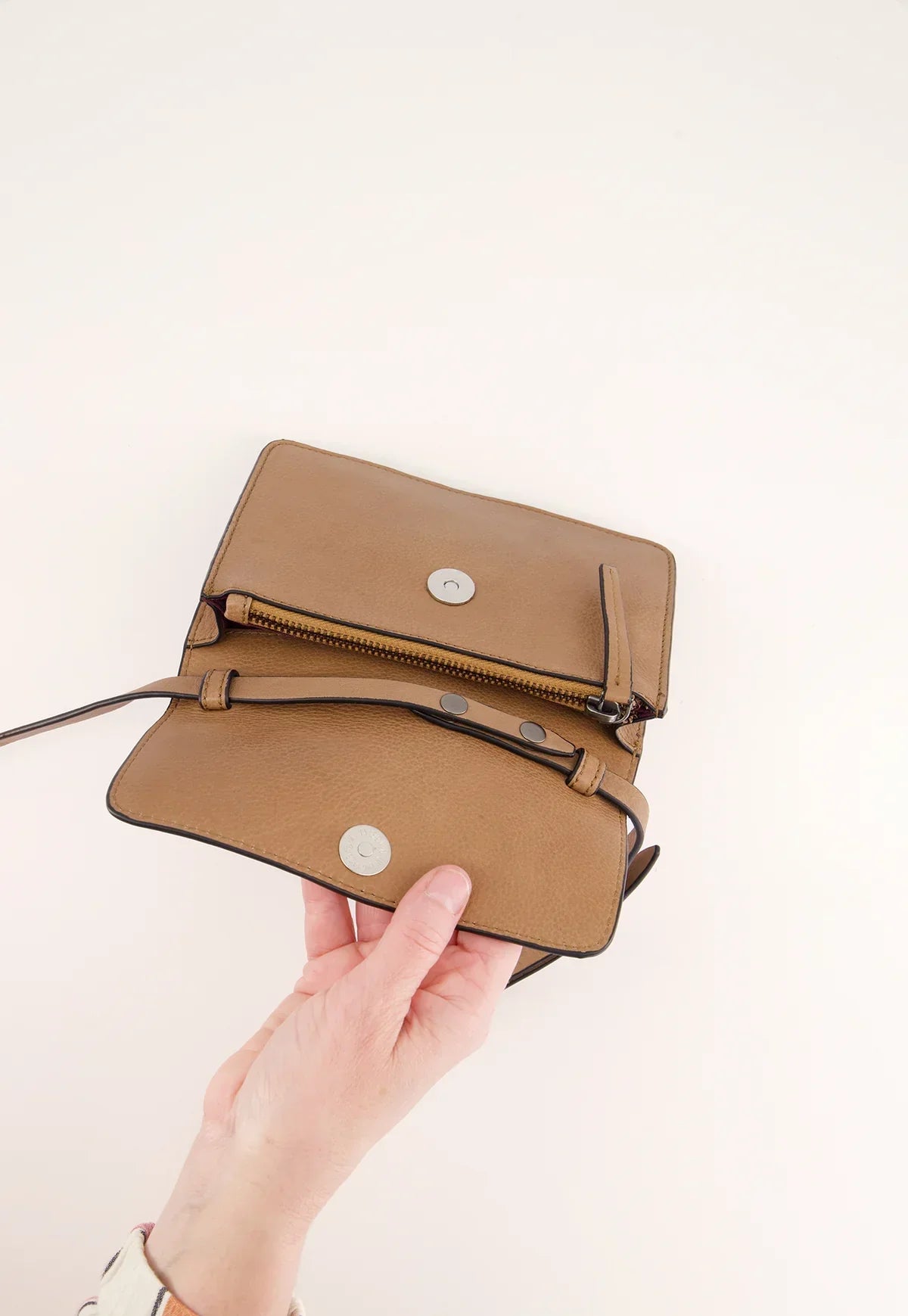 Hand holding open tan leather crossbody bag with magnetic closure and zipper pocket