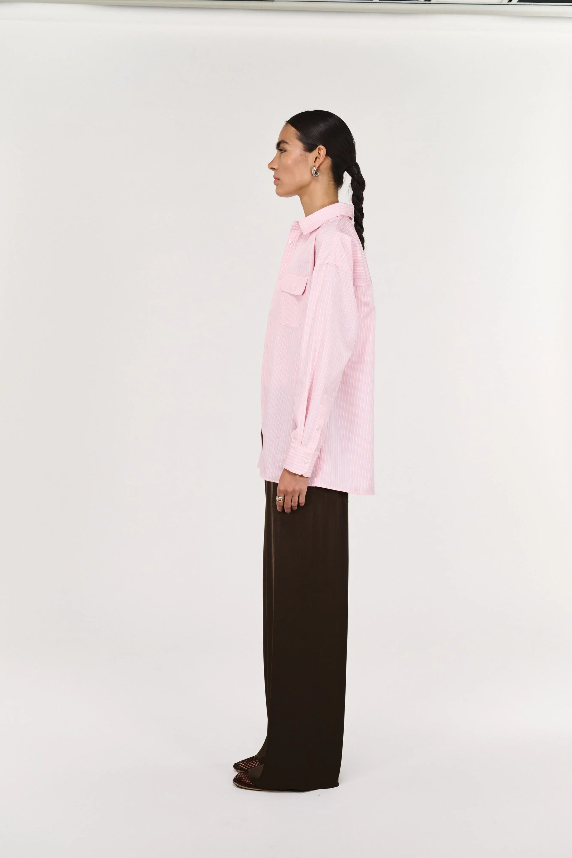 Woman in a pink striped oversized shirt and brown wide-leg pants, side profile, studio background
