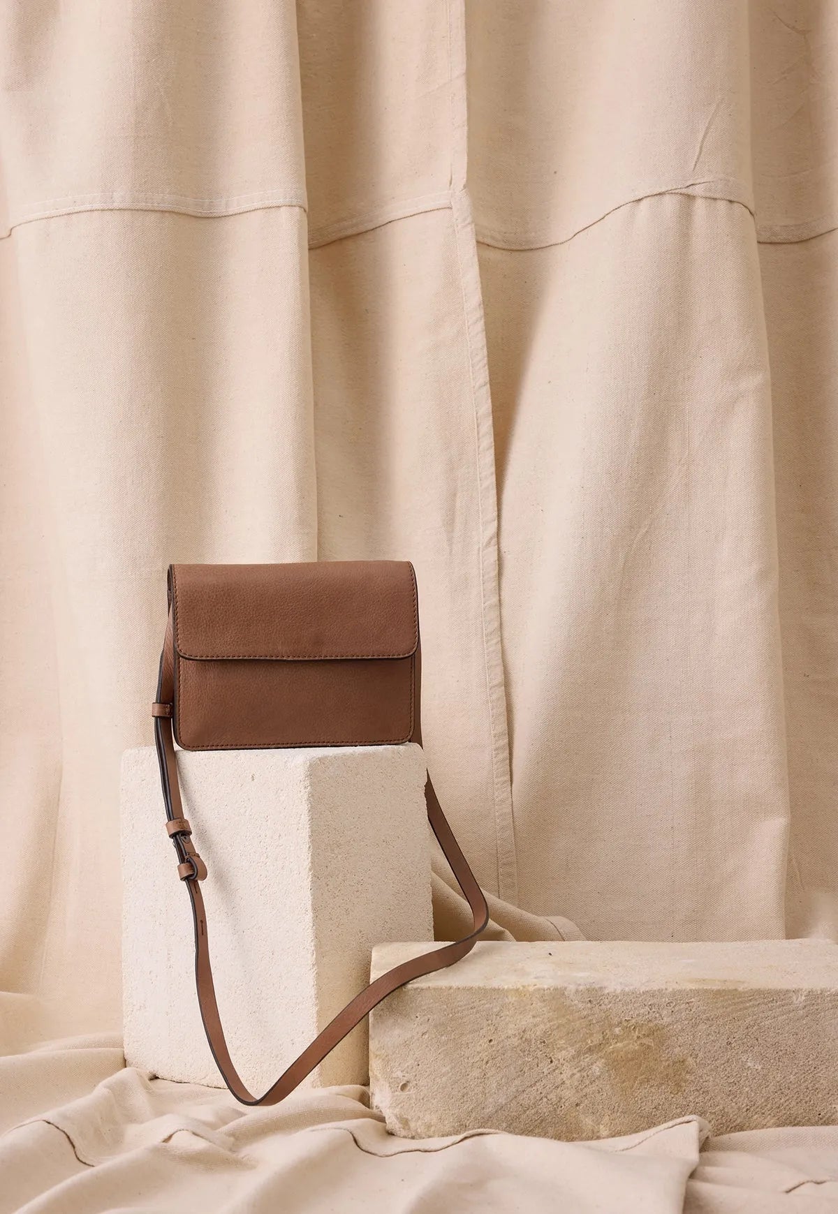 Brown leather crossbody bag with adjustable strap on neutral stone blocks and fabric backdrop