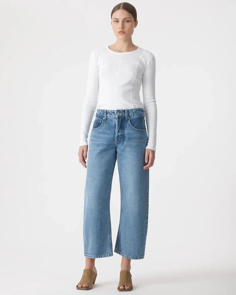 Woman wearing long sleeve white top, wide-leg blue jeans and tan heels, studio background