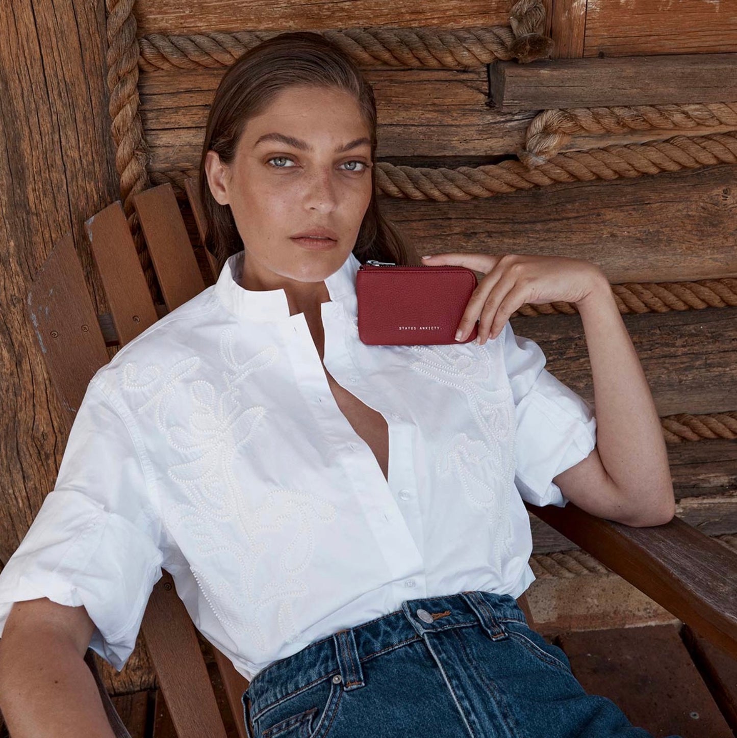 Woman in white embroidered shirt holding red leather wallet, styled with denim jeans against rustic wood background