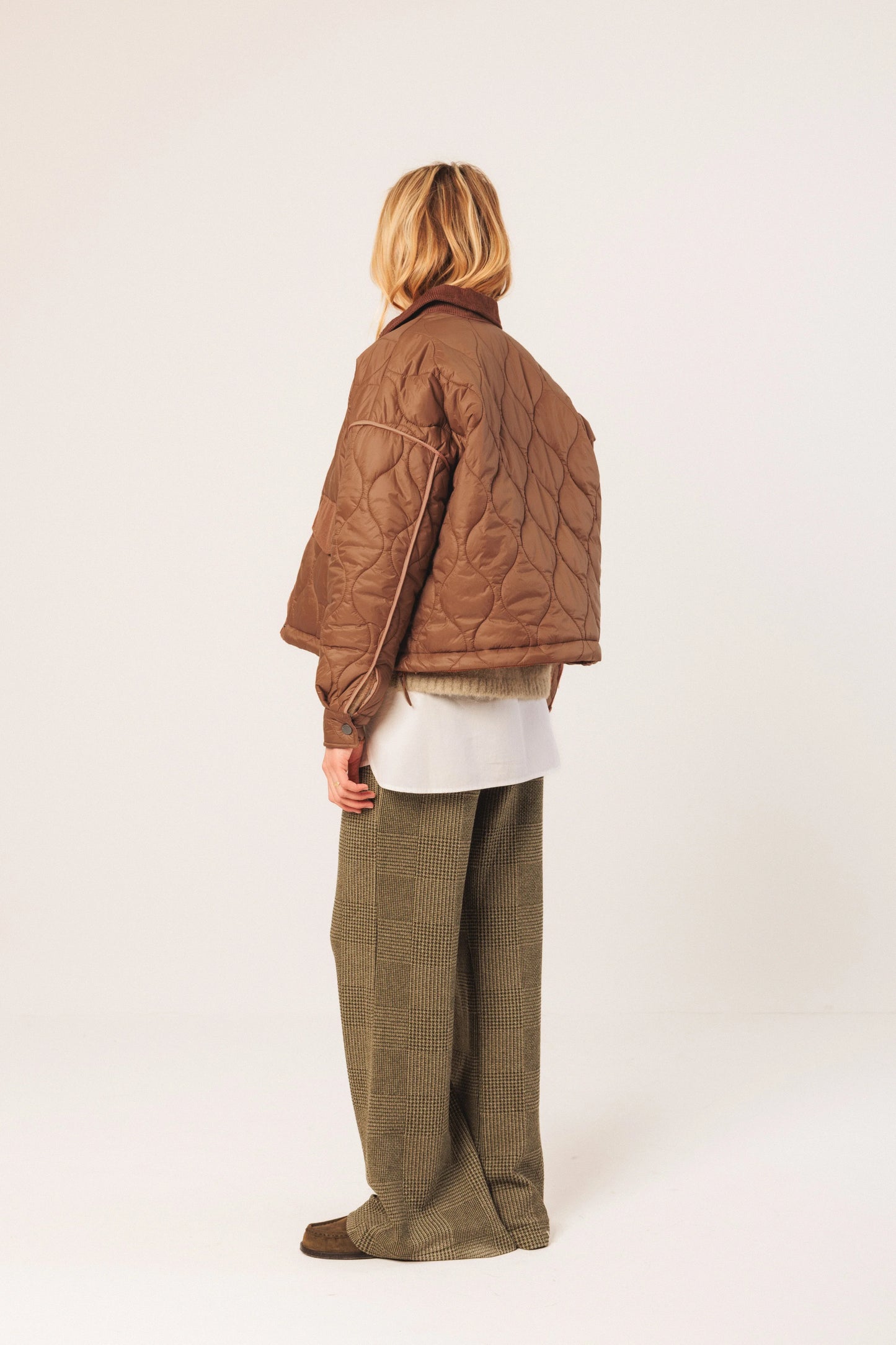 Woman in brown quilted jacket, olive tartan pants, and white shirt, back view