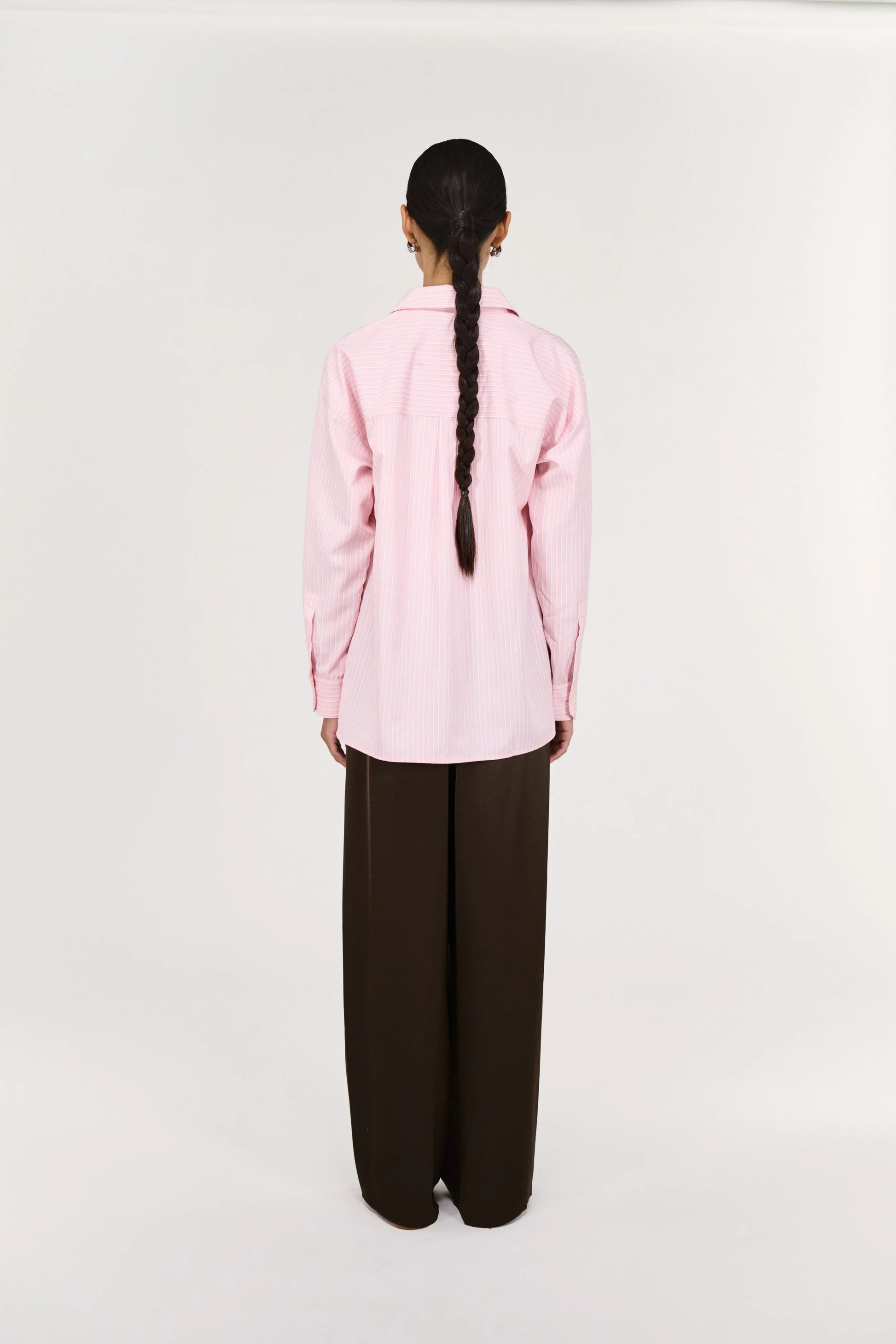 Woman in a pink striped shirt and brown wide-leg pants, standing against a plain background